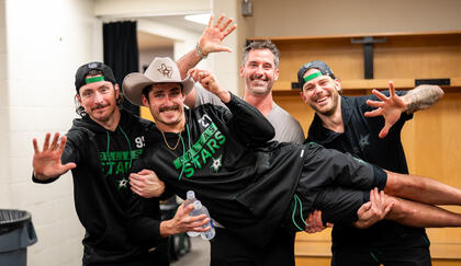 a picture of mason marchment (mush) being held by matt duchene, tyler seguin, and a dallas stars trainer. mush tips a cowboy hat to commemorate a win, and the other men hold up 5 fingers to represent mush's 5-point game.