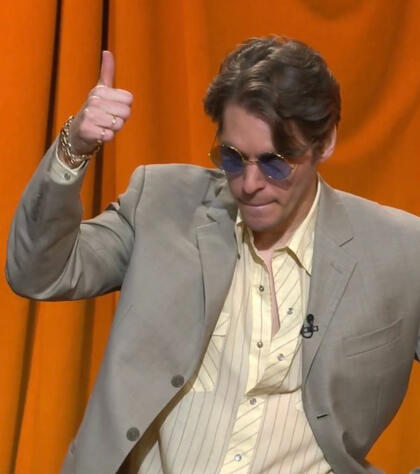 a picture of jerma from the first time he was on name your price. he wears a grey suit and a striped off-white undershirt. he also wears blue-toned sunglasses and he's standing in front of a rich orange curtain. he looks slightly down and gives a thumbs up