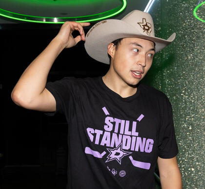 a picture of jason robertson. he wears a black shirt with the dallas stars logo & the text "still standing" in purple. he also wears a beige cowboy hat with the stars logo. he reaches up to the brim & looks off camera.