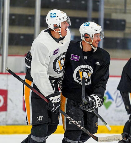 a picture of evgeni malkin and sidney crosby on the ice for practice. geno is leaning on sid and opening his mouth to say something while sid smiles.