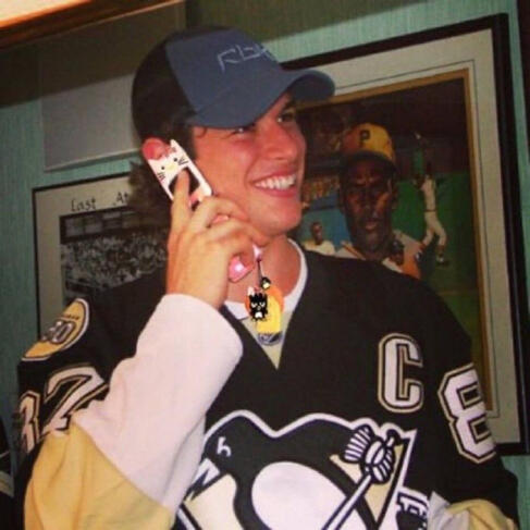 a picture of a young sidney crosby in his black penguins jersey. he's wearing a blue baseball cap, holding a hello kitty flip phone up to his ear, and smiling widely. his face is red.